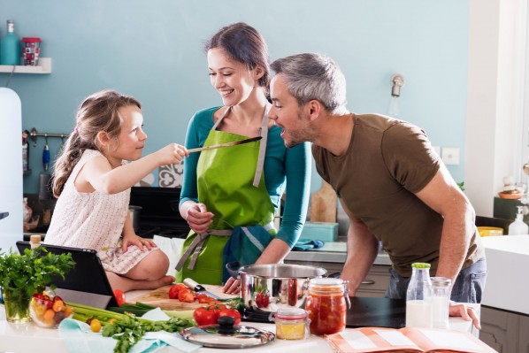 Eine Familie steht in der Küche und kocht gemeinsam. Die kleine Tochter füttert den Vater mit einem großen Holzlöffel.