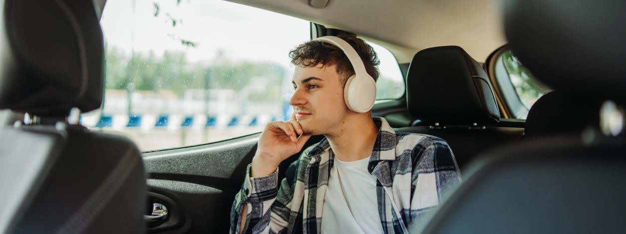 Ein etwa 20-jähriger Mann sitzt hinten im Auto und schaut verträumt aus dem Fenster. Dabei trägt er Kopfhörer.