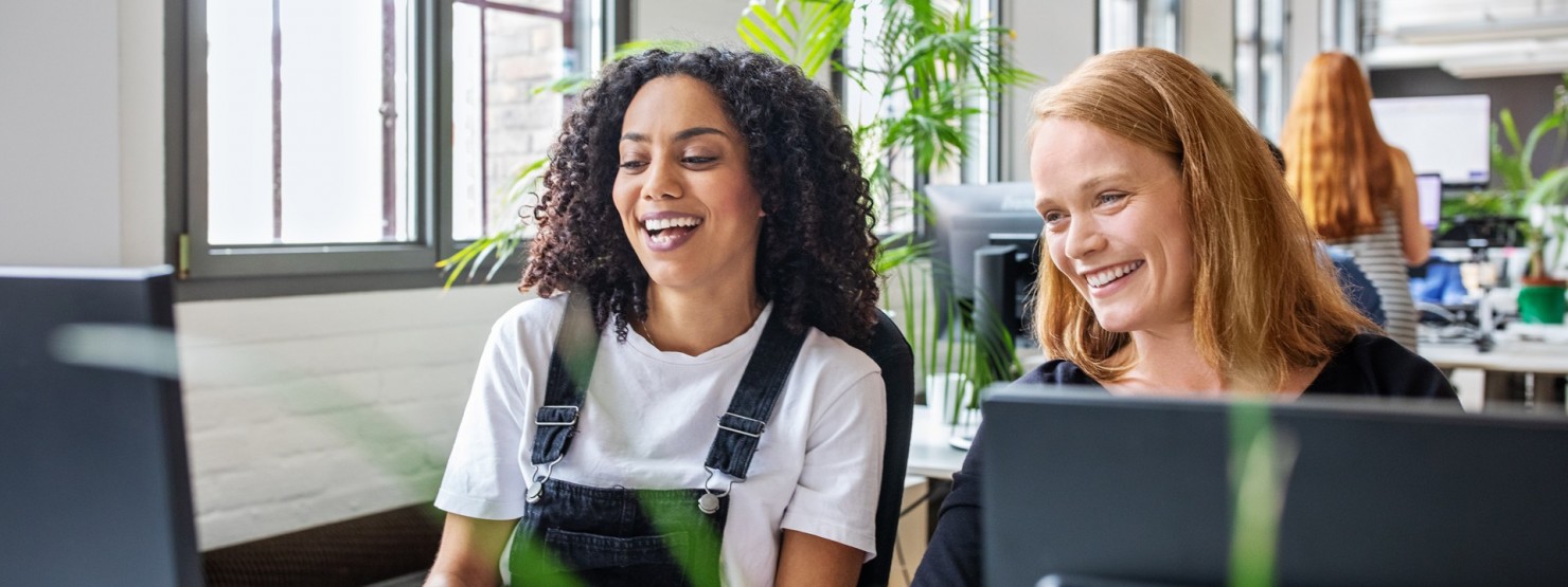 Zwei etwa 30 -jährige Frauen sitzen lachend vor einem Computer am Schreibtisch in einem Großraumbüro.