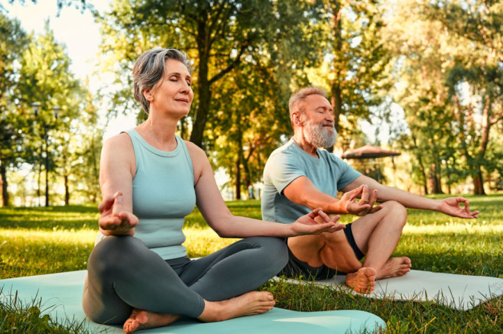 Ein Paar sitzt in einem Park auf ihren Yogamatten und machen Yoga mit der AOK Yogaschule.