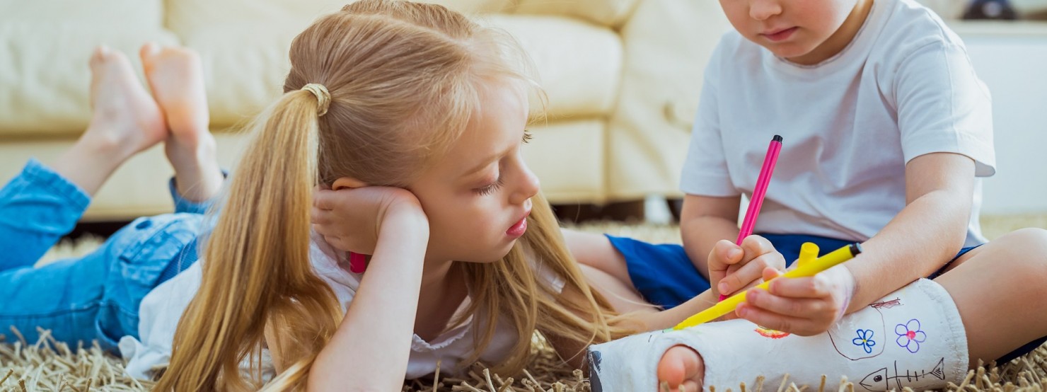 Zwei Kinder liegen auf dem Boden und bemalen einen Fußgips.