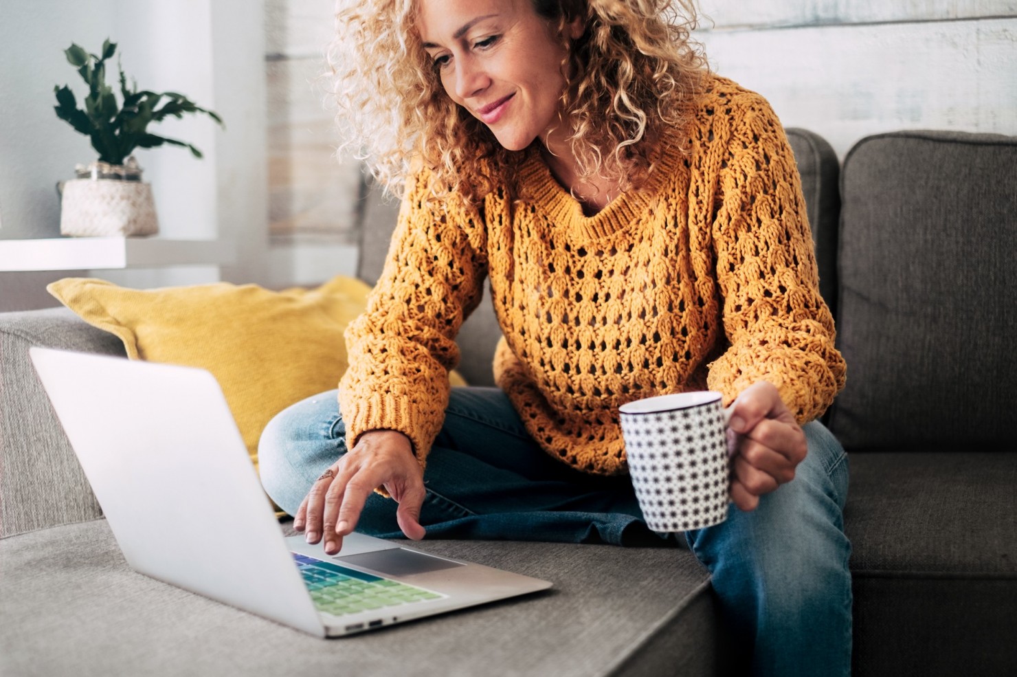 Eine etwa 40-jährige Frau sitzt mit dem Laptop auf dem Sofa. Sie hält eine Tasse in der linken Hand.