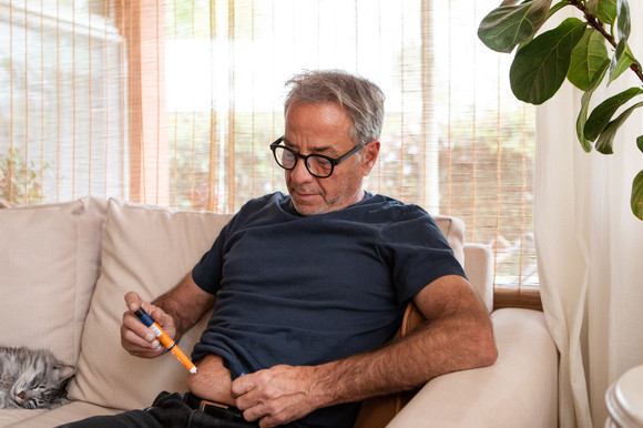Ein etwa 55-jähriger Mann sitzt auf dem Sofa im Wohnzimmer und spritzt sich mit einem Fertigpen in die Bauchfalte.