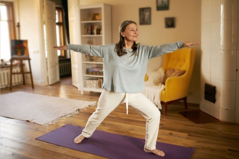 Eine etwa 60jährige Frau steht in einem Wohnzimmer auf einer Yogamatte und macht eine Übung mit ausgestreckten Armen.