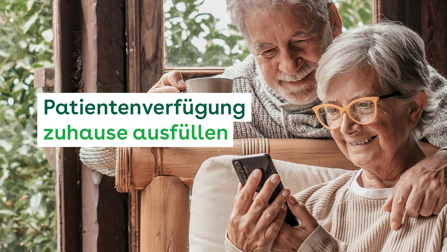 Ein Paar, beide 75 Jahre, sind im Wintergarten und schauen auf das Handy, das die Frau in den Händen hält. Er lehnt sich von hinten über ihre Schulter und hält eine Tasse in der rechten Hand.