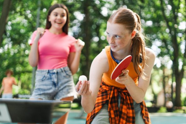 Ein Mädchen spielt Tischtennis. Eine Freundin feuert sie im Hintergrund an.