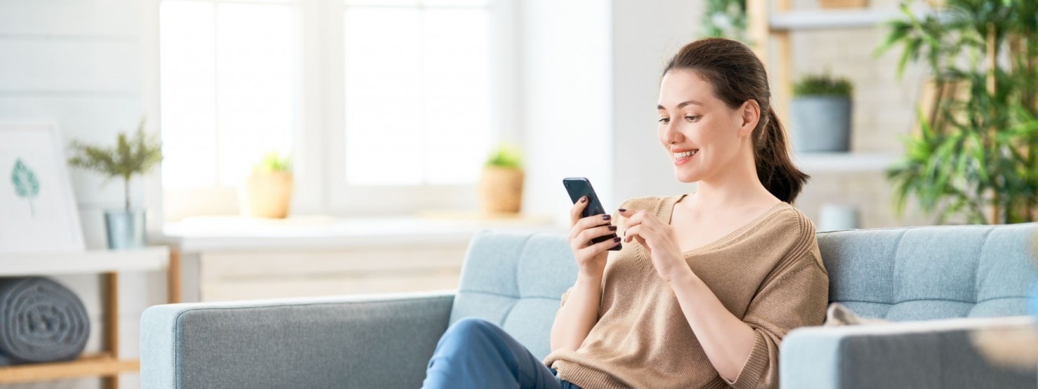 Eine etwas 30 jährige Frau sitzt auf der Couch hat ein Handy in der Hand. Sie lächelt.