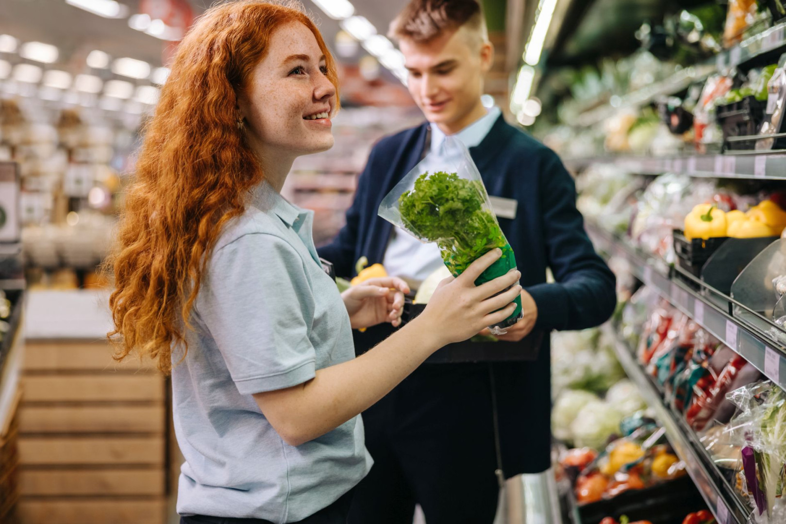 Zwei junge Aushilfen räumen gemeinsam das Gemüseregal in einem Supermarkt ein.