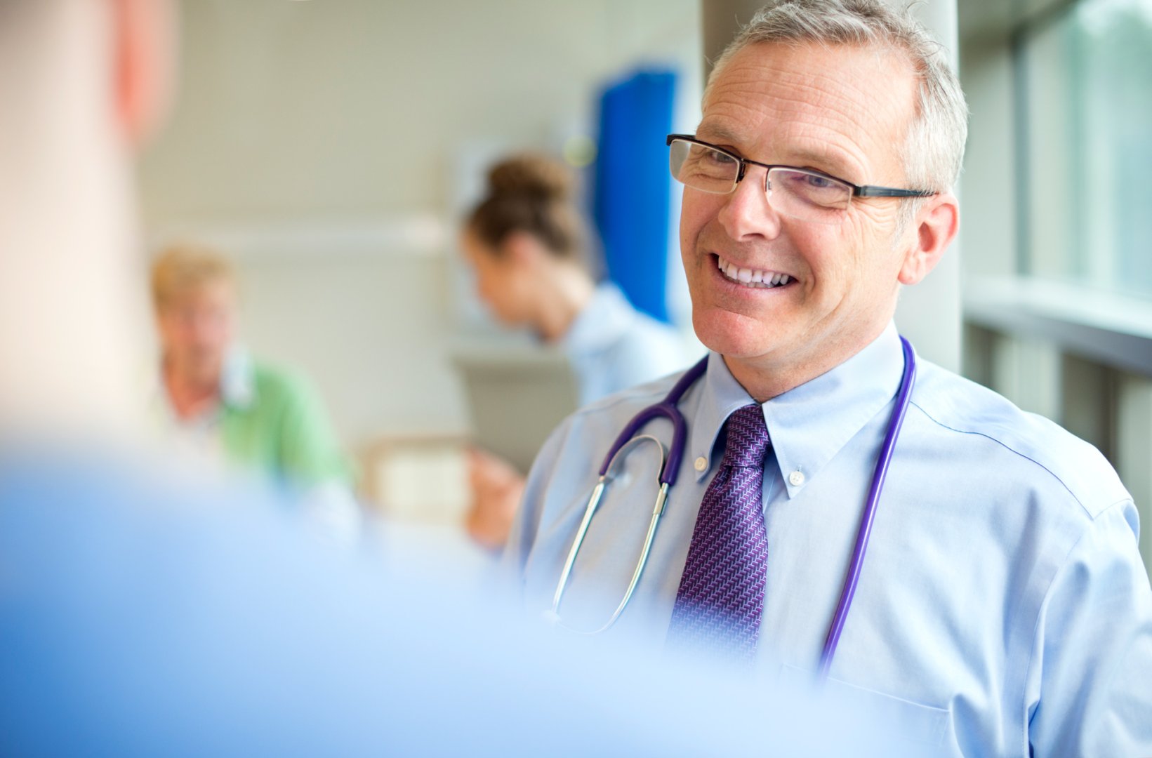 Ein Arzt um die 50 mit Brille steht im Krankenhaus auf dem Flur und führt ein Gespräch mit einem Patienten.