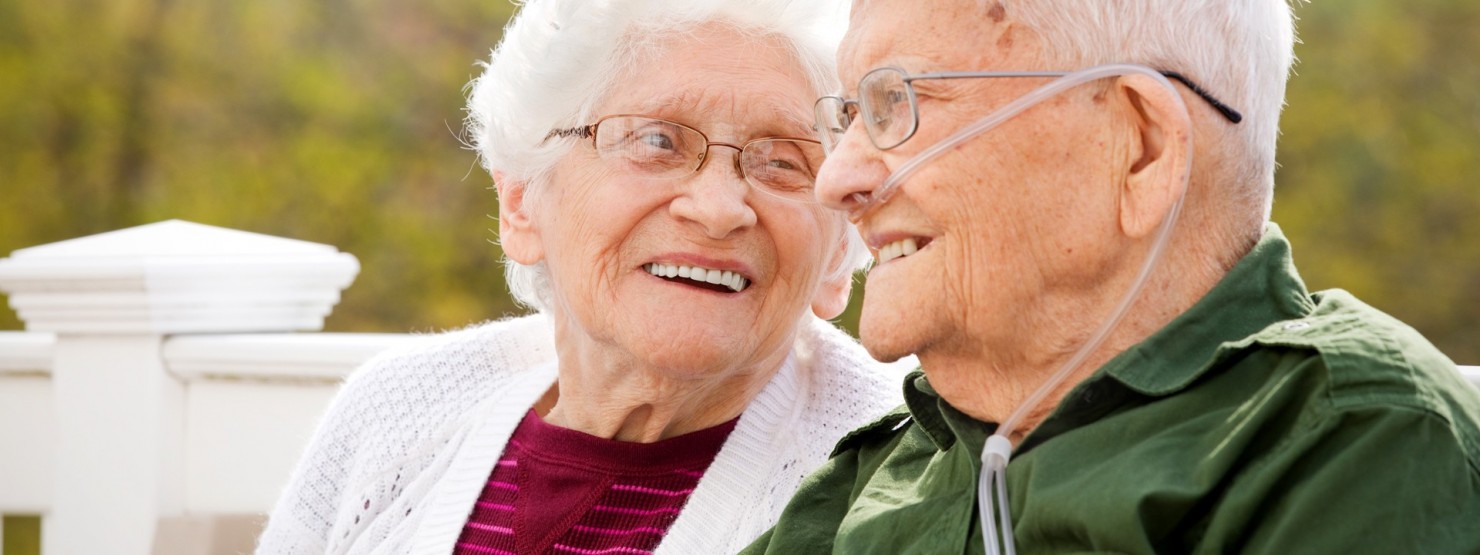 Ein Seniorenpaar sitzt auf einer weißen Bank und schauen sich lächeln an. Der Mann trägt ein Sauerstoffgerät.