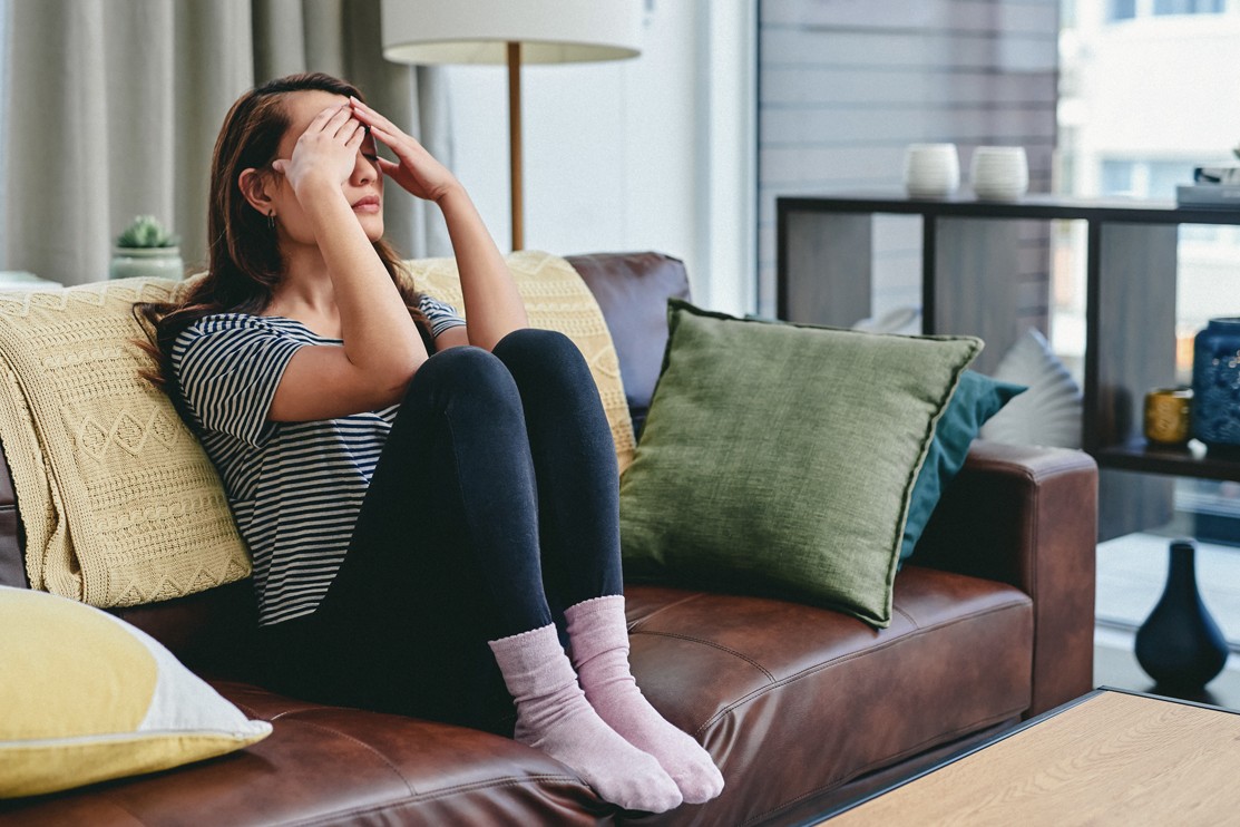 Eine etwa 30-jährige Frau sitzt auf ihrem Sofa und reibt mit beiden Händen Stirn und Schläfen.