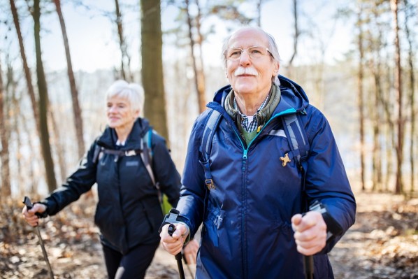 Ein Seniorenpaar läuft mit Nordic-Walking-Stöckern durch den Wald.