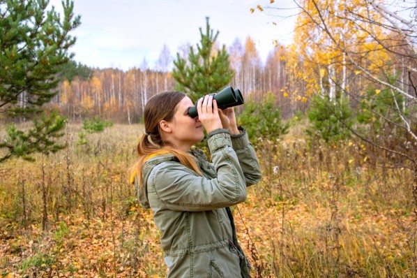 Eine Frau Anfang 30 steht im Herbstwald und beobachtet Tiere durch ein Fernglas.