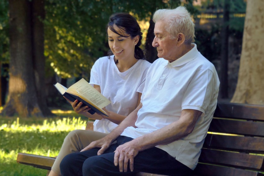 Eine Altenpflegerin sitzt mit einem Senior auf einer Bank in der Natur. Sie liest ihm etwas in einem Buch vor und beide lächeln.