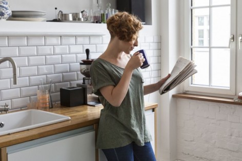 Eine Frau steht in einer Küche, trinkt aus einer Tasse und liest in einer Zeitung.