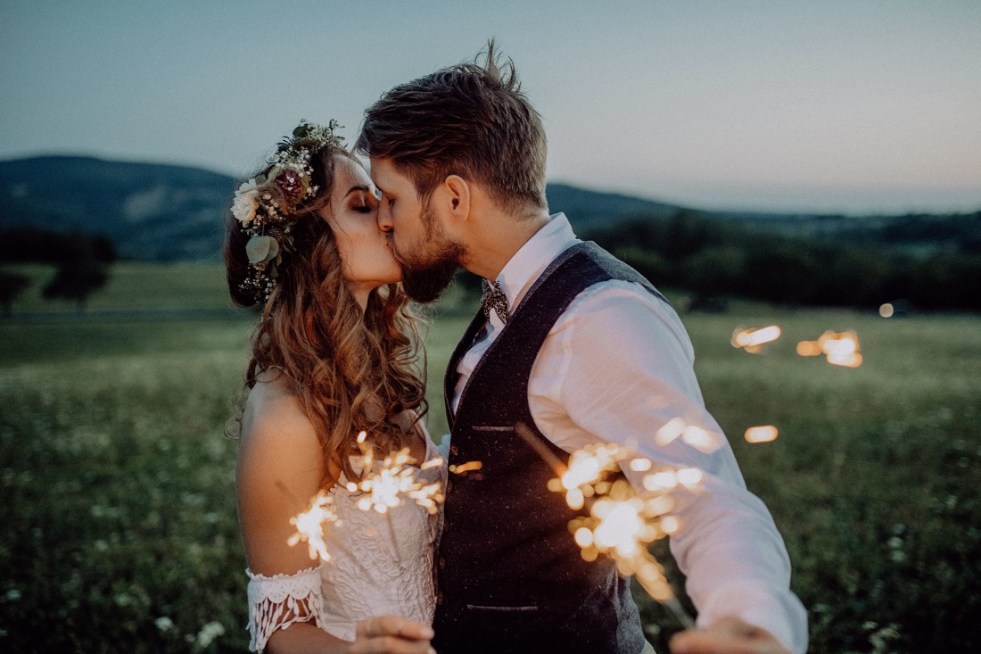 Ein Paar, Mitte 20, steht Abends auf einer Wiese und küsst sich. Sie trägt ein Brautkleid mit Blumenkranz im Haar, er trägt ein weißes Hemd mit schwarzer Weste und Fliege. Sie halten Wunderkerzen. Im Hintergrund sind die Ausläufer eines Mittelgebirges zu sehen.