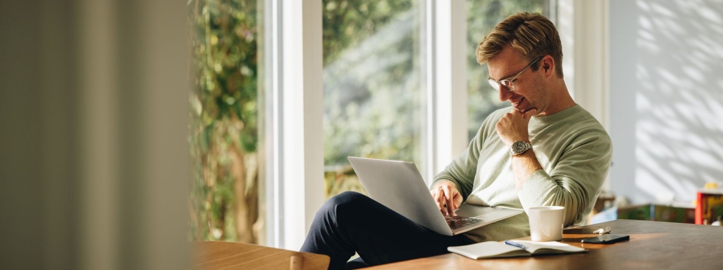 Young man using laptop and smiling at home. Man sitting by table working on laptop computer.