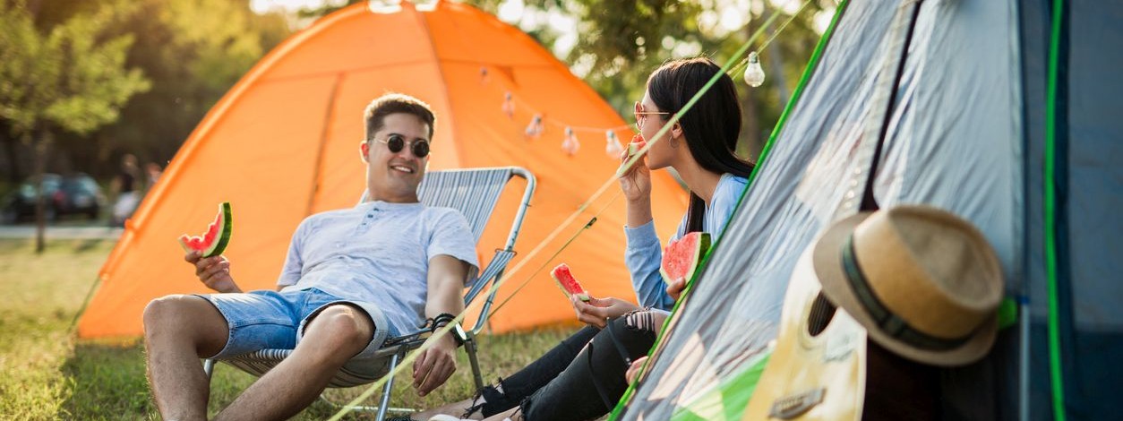 Ein junges Pärchen sitzt vor ihrem Zelt auf einem Festival- Sie essen eine frische Wassermelone. 