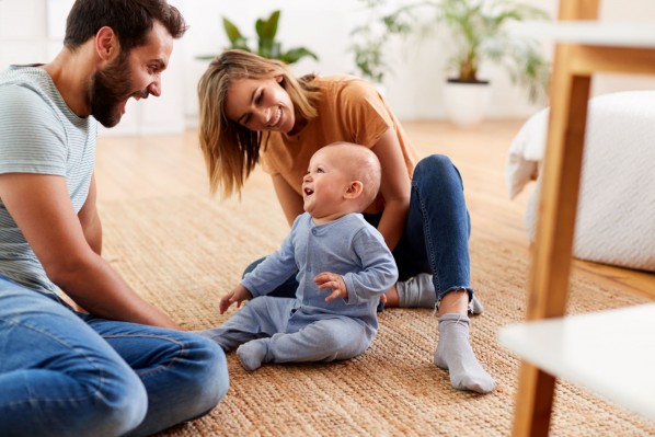 Eine junge Familie sitzt mit ihrem etwa 4 Monate alten Baby auf dem Boden. Die Mutter stützt das Kind beim Sitzen. Alle lachen sich gegenseitig an.