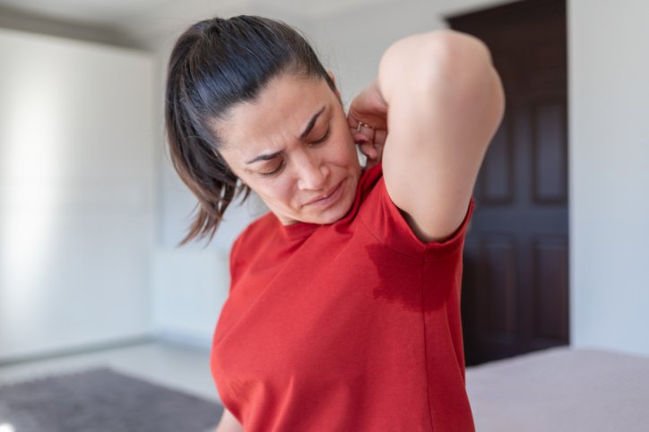 Eine Frau hebt ihren linken Arm. An ihrem T-Shirt zeichnet sich unter der Achsel ein Schweißfleck ab. 