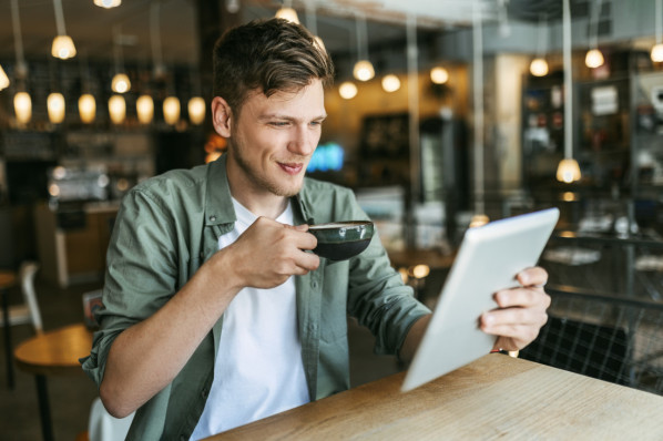 Ein etwa 30-jähriger Mann sitzt in einem Café und schaut auf ein Tablet.