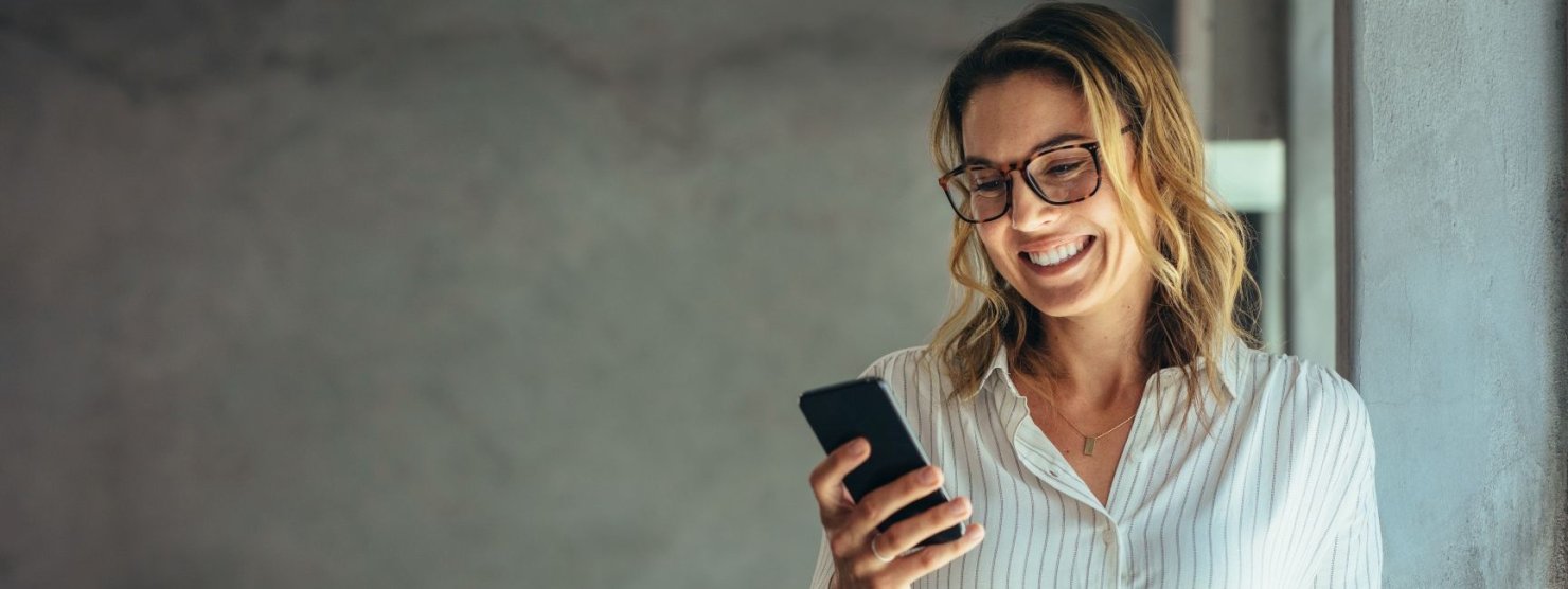 Eine etwa 40 jährige Frau steht an einem Fenster und schaut lächelnd auf ihr Handy.