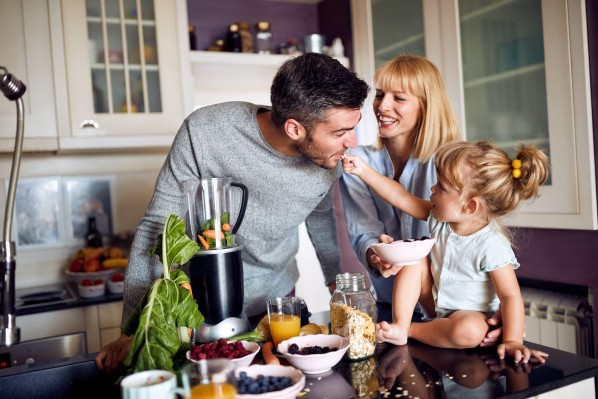 Eine dreiköpfige Familie steht in der Küche und macht Frühstück. das kleine Mädchen sitzt auf dem Küchentresen und füttert den Vater.