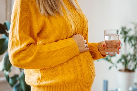 Eine Frau hält mit der einen Hand ein Wasserglas und mit der anderen ihren Babybauch. 