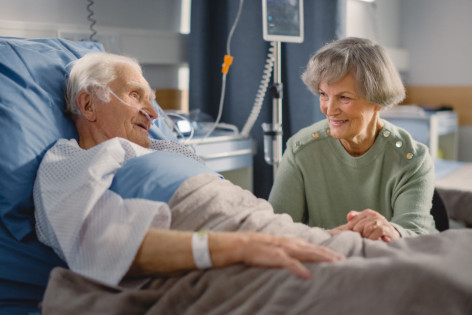 Ein älterer Patient liegt in einem Krankenhausbett und hat Besuch von einer Angehörigen. Er nimmt den Qualitätsvertrag zum postoperativen Delir in Anspruch.