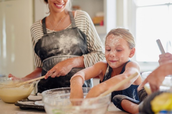 Ein etwa 6-jähriges Mädchen steht neben ihrer Mutter in der Küche und fasst beherzt mit beiden Händen in die Teigschüssel. Beide sind gut gelaunt und im Gesicht und an den Schürzen mit reichlich Mehl bestäubt.