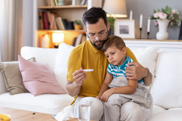 A concerned dad holds a thermometer while his sick child rests on his lap on a white couch, depicting attentive and loving parental care in a comfortable home setting.