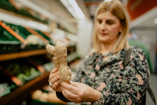 Eine Frau steht in einem Supermarkt am gemüseregal und schaut sich eine große Ingwerknolle an.