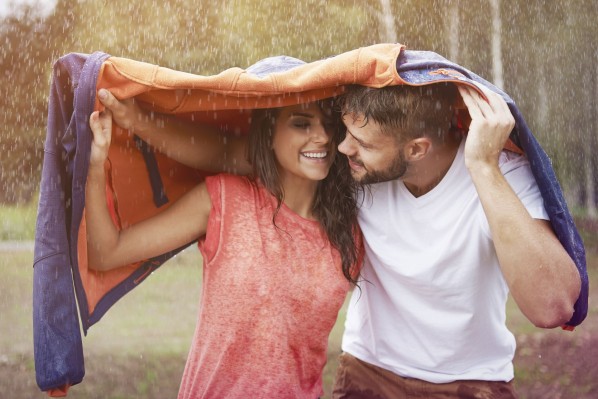 Ein Paar läuft durch strömenden Regen. Als Regenschutz haben sie eine Jacke über ihre Köpfe geworfen. Beide schauen sich lächelnd an.