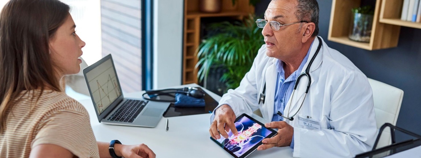 Eine Patientin und ein Orthopäde besprechen sich in einem Arztzimmer. Der Orthopäde zeigt der Patientin etwas auf seinem Tablet.