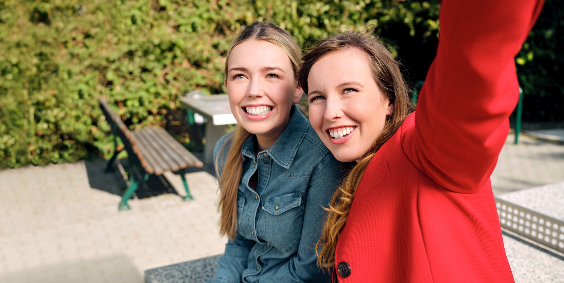 Die Auszubildenden Alina und Josi sitzen in einem Park auf einer Tischtennisplatt und machen gemeinsam ein Selfie.
