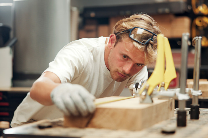 Ein etwa 19-jähriger Zimmermann bearbeitet ein Stück Holz mit einem Werkzeug. Er blickt konzentriert auf seine Arbeit. Er trägt eine Schutzbrille auf dem Kopf und Arbeitshandschuhe.