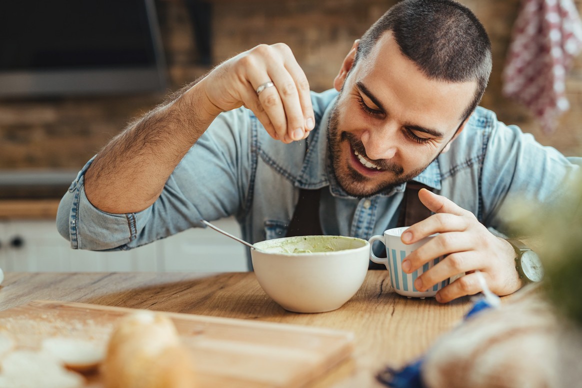 Ein etwa 30-jähriger Mann würzt lächelnd eine Schüssel Suppe.