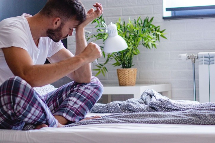 Ein Mann sitzt auf dem Bett und hält sich den Kopf.