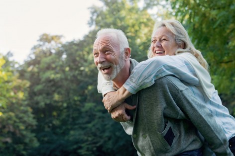 Ein Seniorenpaar hat gemeinsam in der Natur Spaß. Der Mann trägt die Frau auf dem Rücken. Beide lachen ausgiebig.