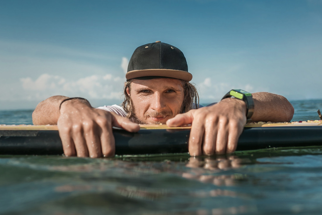 Junger Mann mit Cap lehnt entspannt auf Surfbrett im Meer – AOK Sachsen-Anhalt unterstützt junge Menschen in allen Lebenslagen.