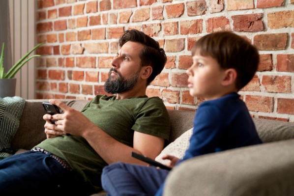 Ein Vater sitzt mit seinem etwa 5 jährigen Sohn auf der Couch. Der Junge hat eine Fernbedienung in der Hand, der Vater sein Handy. Beide schauen interessiert in eine Richtung.