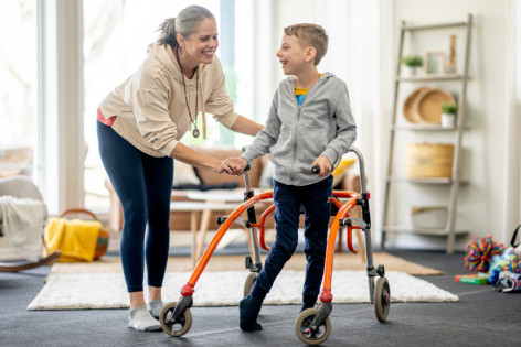 Ein Junge mit Gehbehinderung steht in einer medizinischen Stehhilfe. Seine Mutter unterstützt ihn zusätzlich.