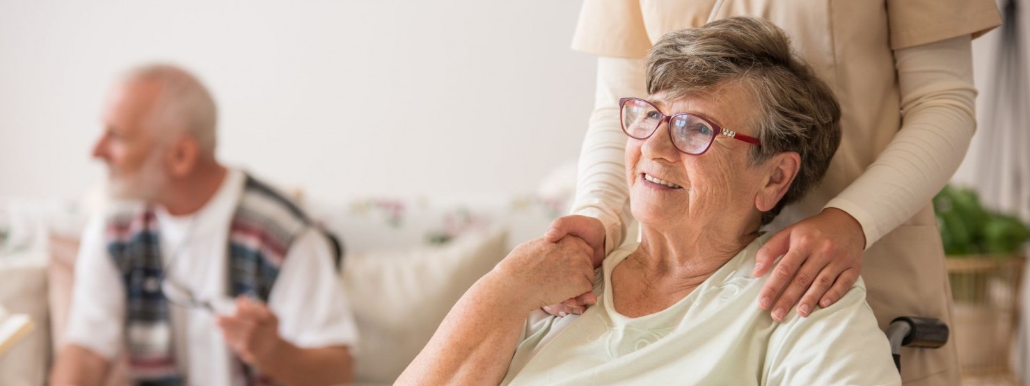 Eine Seniorin sitzt in einem Rollstuhl. Eine weibliche Person fasst ihr von hinten an die Schultern. Die Seniorin lächelt.