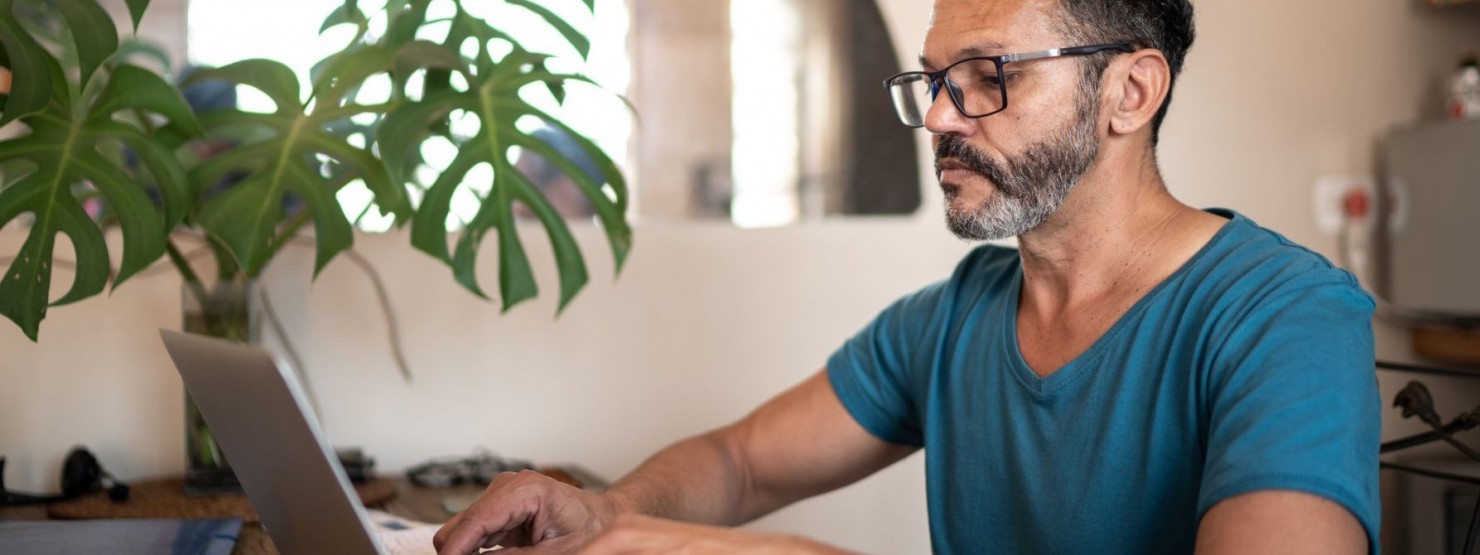 Ein etwa 40 jähriger Mann sitzt an einem Tisch und tippt auf seinem Laptop.