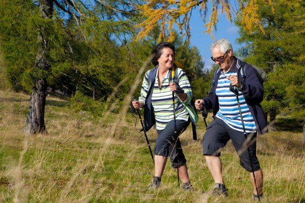 Ein Seniorenpaar betreibt Nordic-Walking in der Natur. Beide lächeln ausgelassen.