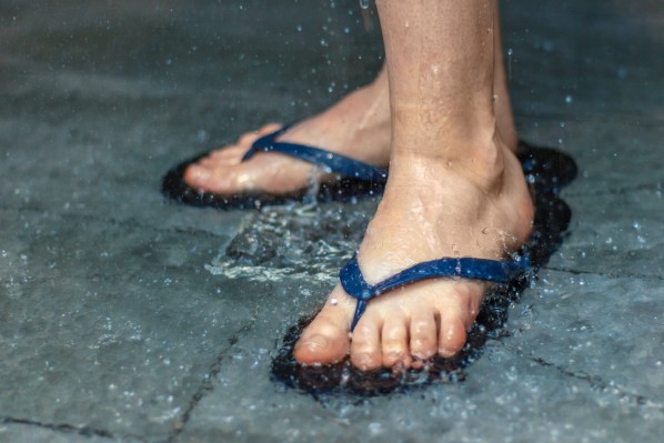 Füße in Badelatschen stehen in einer öffentlichen Dusche. Wasser prasselt auf die Fliesen.