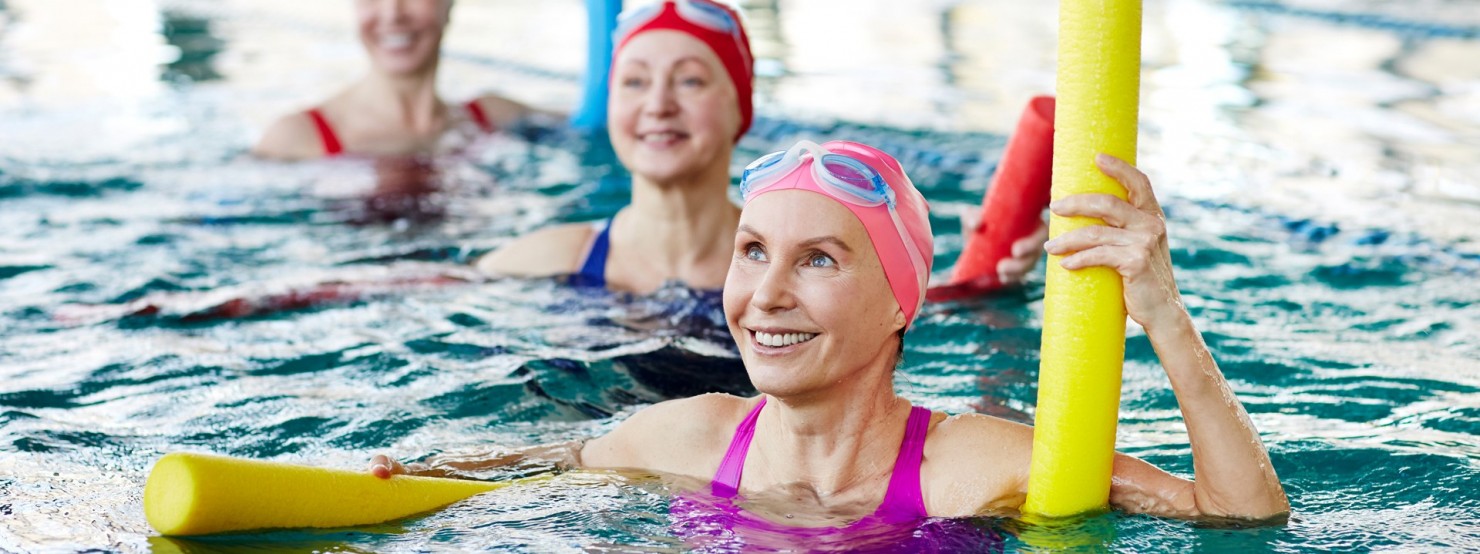 Drei Frauen befinden sich in einer Schwimmhalle im Wasser und machen Wassergymnastik mit bunten Badenudeln.
