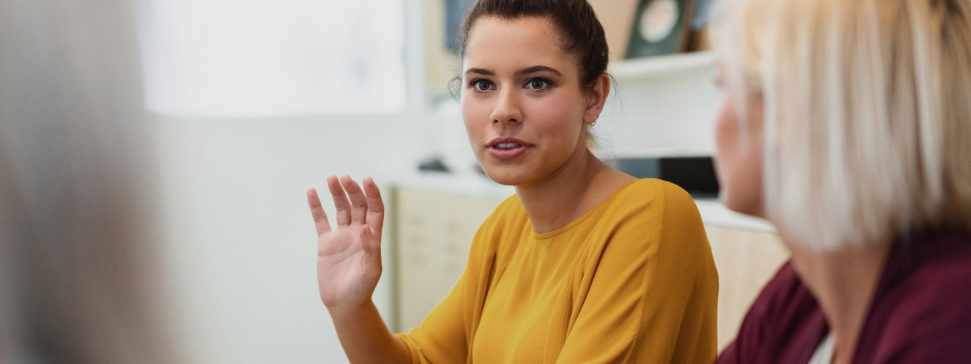 Eine Frau, etwa 30 Jahre, sitzt mit 2 Kolleginnen an einem Tisch. Sie gestikuliert erklärend mit einer Hand.