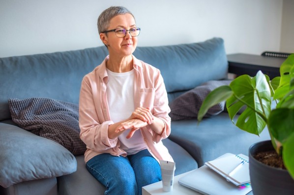 Eine etwa 60-jährige Frau sitzt auf dem Sofa und cremt sich die Hände ein.