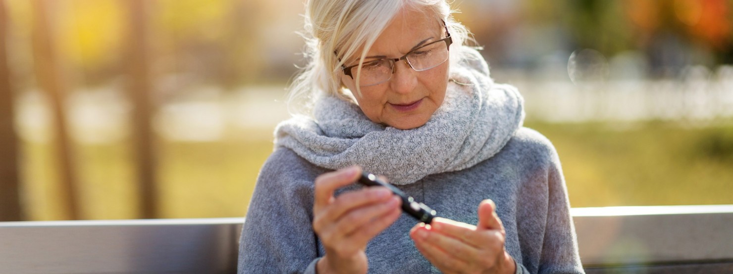 Frau Mitte 60 sitzt im Herbst auf einer Parkbank und misst ihren Blutzuckerspiegel.